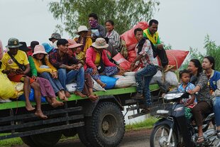Viele Menschen sitzen auf der offenen Ladefläche eines LKW, daneben fährt eine Frau ein Mofa, auf dem auch ihre beiden Töchter sitzen