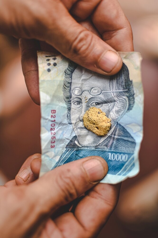 Venezolanischer Geldschein, auf dem ein kleiner Goldklumpen liegt, wird von zwei Händen gehalten