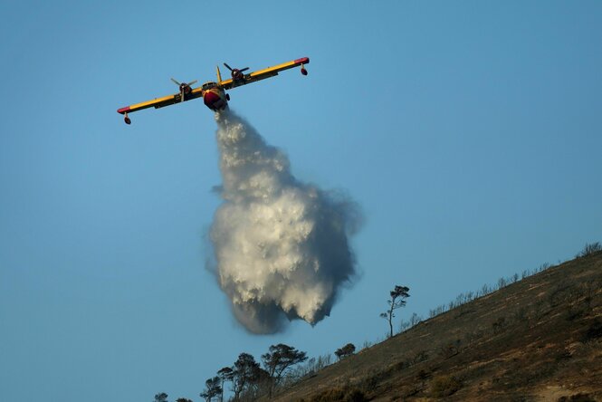 Ein Flugzeug stößt im Flug eine Ladung Wasser aus, unter ihm eine verbrannte Landschaft