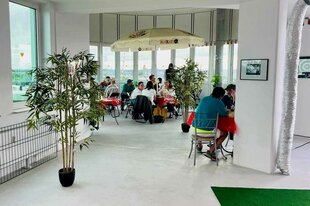 ein heller Raum mit weitem Ausblick, der Vordergrund ist eher leer, im Hintergrund sitzen Menschen an Tischen