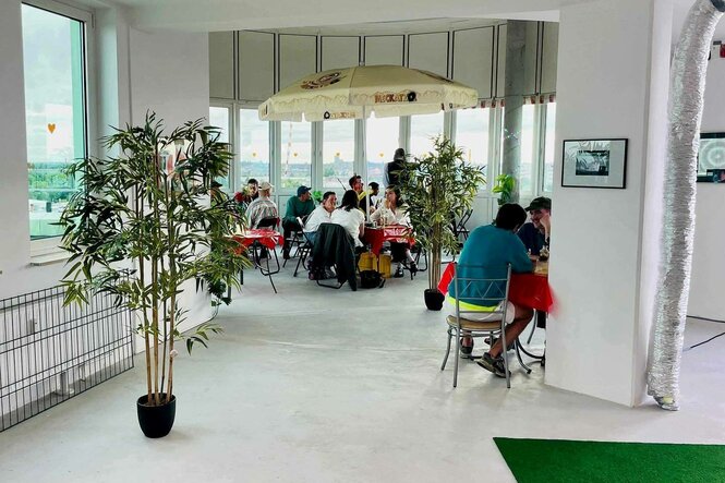 ein heller Raum mit weitem Ausblick, der Vordergrund ist eher leer, im Hintergrund sitzen Menschen an Tischen