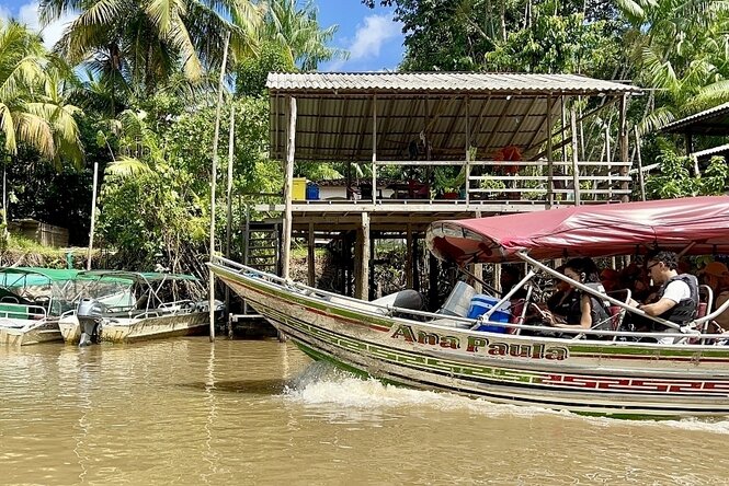Überdachtes Motorboot fährt mit Gästen von rechts durchs Bild. Im Hintergrund ist ein Pflahlbau und angelegte Motorboote zu sehen.