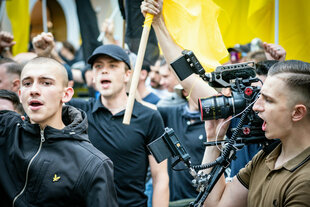 Mehrere Männer in schwarz halten Flaggen der identitären Bewegung. Rechts davon ein Fotograf mit Seitnscheitel im Profil
