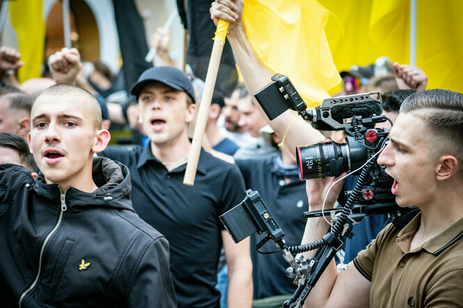 Mehrere Männer in schwarz halten Flaggen der identitären Bewegung. Rechts davon ein Fotograf mit Seitnscheitel im Profil