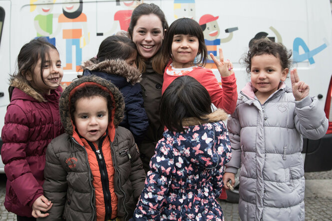 Eine junge Frau steht mit sieben Kindern in bunter Winterkleidung vor einem Fahrzeug mit Comicfiguren darauf. Zwei Kinder zeigen fröhlich das Peace-Zeichen, andere lachen oder schauen neugierig. Die Kinder tragen dicke Jacken, einige mit Fellkapuze, und wirken vertraut mit der Frau in ihrer Mitte.