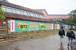 Passanten gehen im Regen an der mit Plakaten beklebten Bretterwand vor dem Sport- und Erholungszentrum (SEZ) an der Landsberger Allee vorbei.