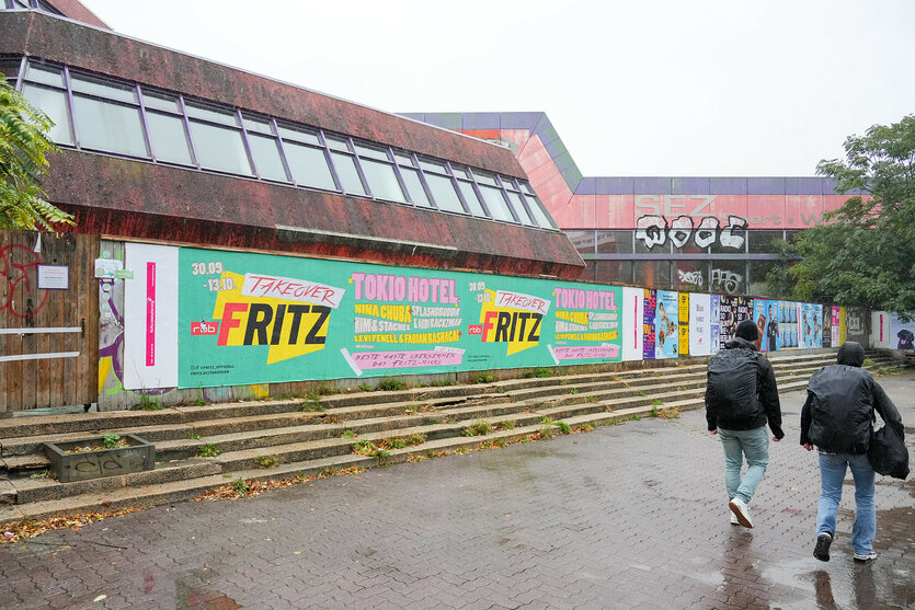 Passanten gehen im Regen an der mit Plakaten beklebten Bretterwand vor dem Sport- und Erholungszentrum (SEZ) an der Landsberger Allee vorbei.