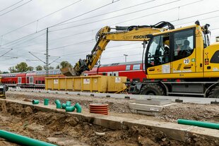 Ein Bagger bei Erdarbeiten entlang einer Bahntrasse, im Hintergrund ein Regionalzug