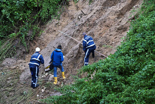 Drei Männer in blauer Arbeitskleidung stehen an dem abgerutschten Hang und vermessen die Stelle
