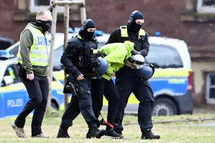 Der Tatverdächtige der Messerattacke am Holocaust-Mahnmal wird von vier vermummten Männern zu einem Hubschrauber geführt.