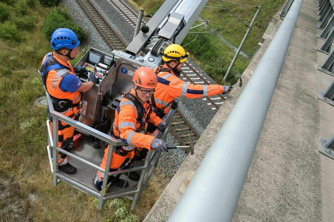 Drei Männer stehen in orangefarbener Arbeitskleidung in einem Krankorb und arbeiten an einer Brücke