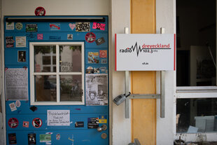 Aussenansicht der Redaktion Radio Dreyeckland - eine blaue Tür mit vielen bunten Stickern und Plakaten und einem Schild mit dem Logo vom Radio