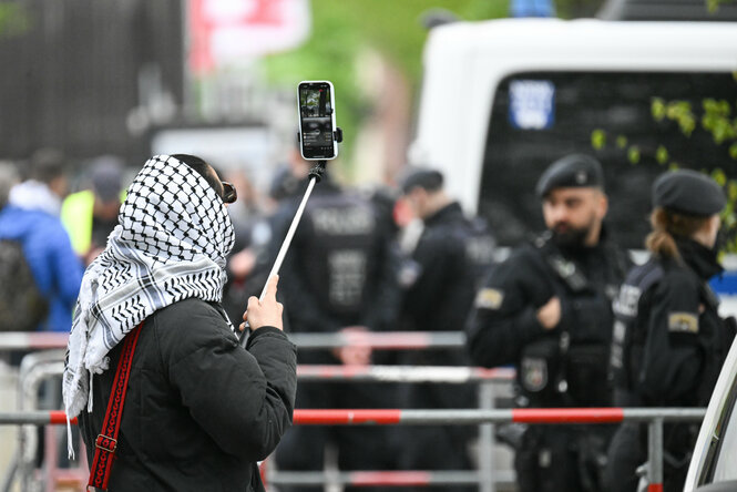 Ein Mensch mit Kufiya filmt Polizisten hinter einer Absperrung