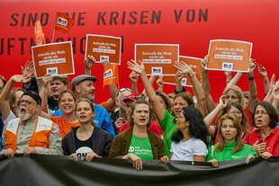 Eine Gruppe von Menschen unterschiedlichen Alters steht vor einer roten Wand und hält Protestschilder hoch
