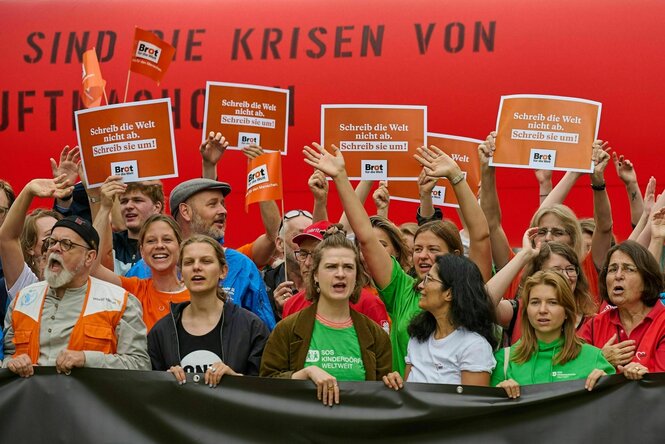 Eine Gruppe von Menschen unterschiedlichen Alters steht vor einer roten Wand und hält Protestschilder hoch