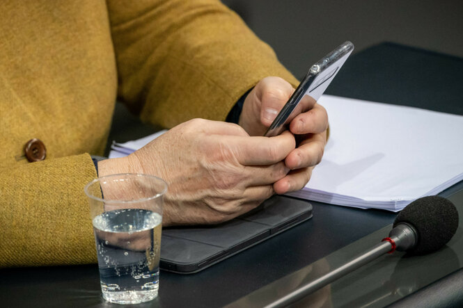 Nahaufnahmer einer Person im senfgelben Jackett hält ein Smartphone in beiden Händen. Auf dem Tisch liegen Unterlagen, ein Tablet, ein Mikrofon – und ein Plastikbecher mit Sprudelwasser. Kontext: Die Nahaufnahme zeigt Angela Merkels Hände im Bundestag