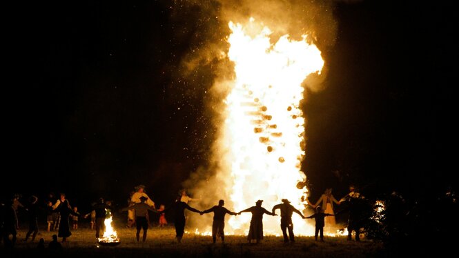 Ein großes Feuer brennt in der Dunkelheit, Menschen halten sich an den Händen und tanzen um die Flammen