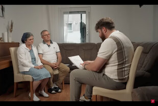 Screenshot: Ein Paar sitzt auf Stühlen - rechts ein Mann, der sie interviewt