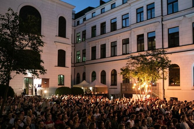Ein bestuhlter Platz, umgeben von alten Gebäuden, an einem sommerlichen Abend. Das Publikum schaut gebannt auf eine Leinwand.