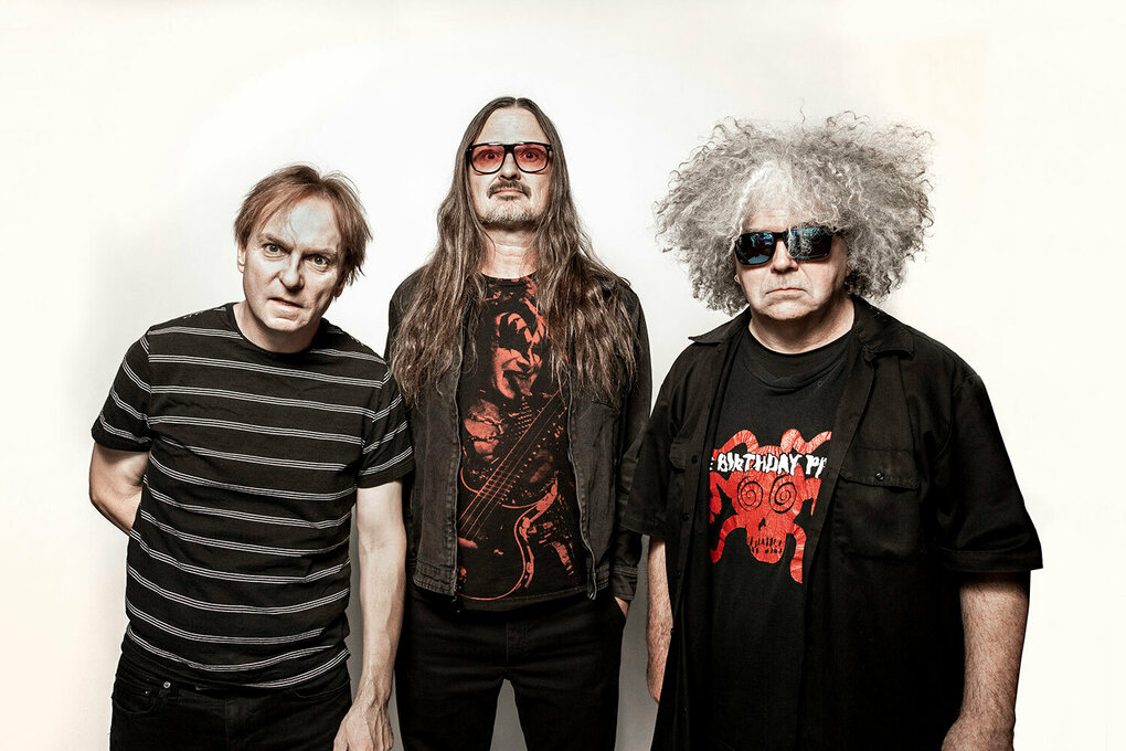 Die drei Mitglieder der Band Melvins stehen vor einem weißen Hintergrund. Sie tragen (von links nach rechts) Ringel-T-Shirt (Dale Crover), getönte Brille (Steven McDonald) und graue Locken (King Buzzo).