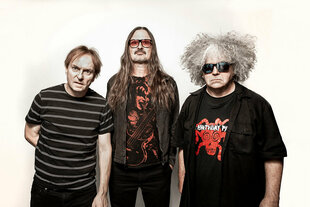 Die drei Mitglieder der Band Melvins stehen vor einem weißen Hintergrund. Sie tragen (von links nach rechts) Ringel-T-Shirt (Dale Crover), getönte Brille (Steven McDonald) und graue Locken (King Buzzo).