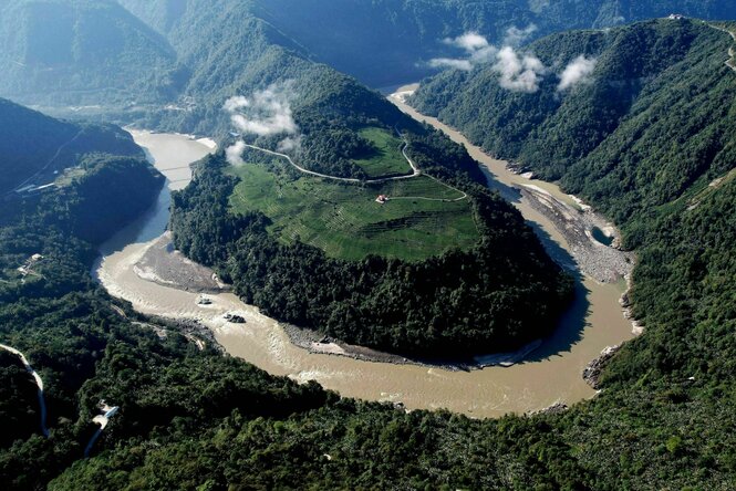 Luftaufnahme des Flusses Yarlung Tsangpo, der eine Schleife um einen großen bewaldeten Hügel macht, im Hintergrund bewaldete Bergketten