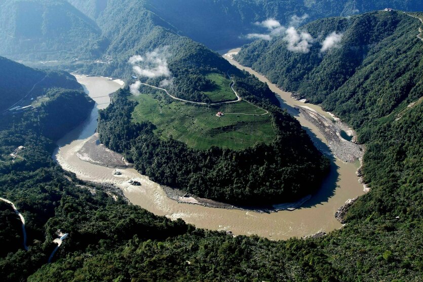 Luftaufnahme des Flusses Yarlung Tsangpo, der eine Schleife um einen großen bewaldeten Hügel macht, im Hintergrund bewaldete Bergketten