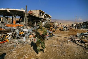 Ein vermummter Soldat geht mit einem Gewehr in der Hand durch Sinjar, das völlig zerstärt ist, die Straßen voll mit Einrichtungsgegenständen, Hauswände fehlen