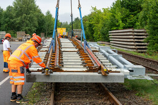 Arbeiter heben mit Hilfe eines Eisenbahndrehkrans eine fertig montierte Weiche zum Transport vom Baumaterial-Lager für den Ausbau der Bahnstrecke Berlin - Hamburg zu einem nahe gelegenen anderen Lagerplatz.