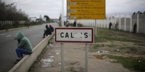 Migranten sitzen am Fahrbahnrand neben einem Ortsschild von Calais.