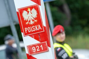 Im Vordergrund markiert ein weiß-roter Pfahl die deutsch-polnische Grenze, im Hintergrund ist eine Grenzbeamte zu sehen