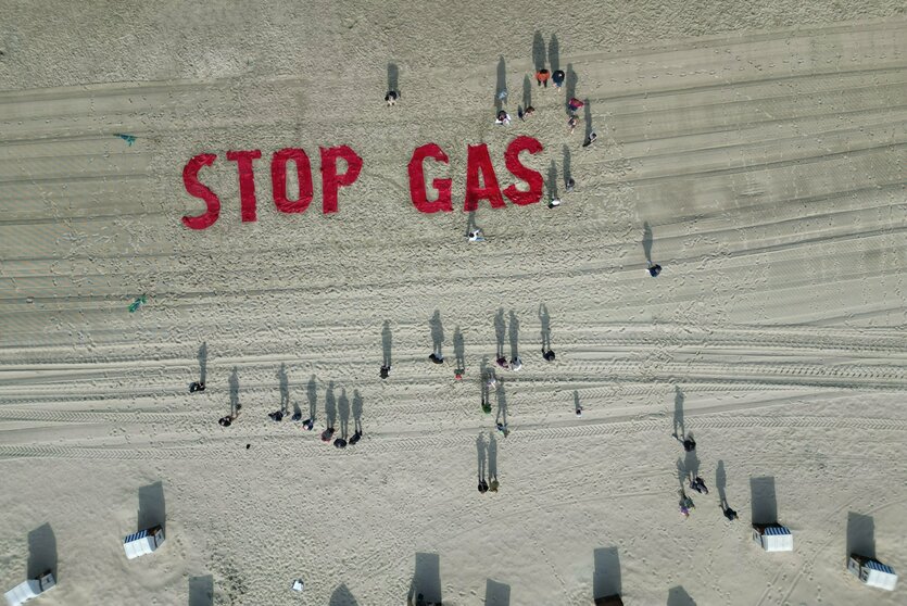Roter Schriftzug "Stop Gas" auf weißem Sand, dazu Schatten von Menschen und Strandkörben