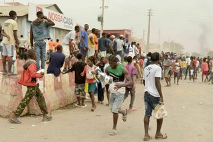 Viele junge Männer stehen auf einer staubigen Strße in Luanda, manche stehen auf einer Mauer. In weiterer Entfernung ist eine Rauchsäule zu sehen