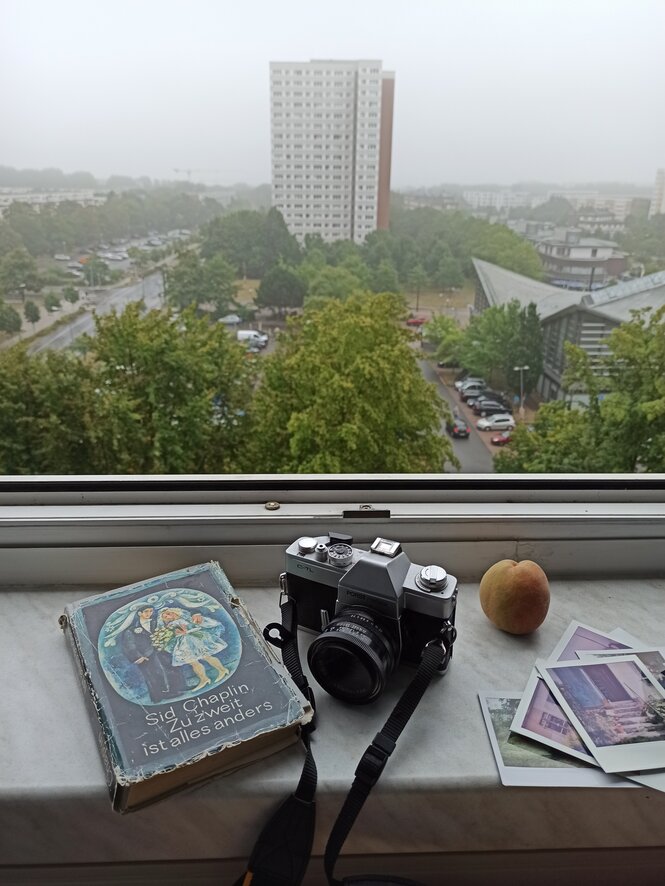 Auf einem Fensterbrett liegen Gegenstände, draußen blickt man auf ein Hochhaus, es ist bewölkt