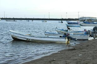 Einfache Boote mit Außenbordmotor liegen am Strand von Obock, in manchen Booten sitzt jeweils ein Mann, im Hintergrund ankern weitere Boote, die Hafenmauer begrenzt das Meer