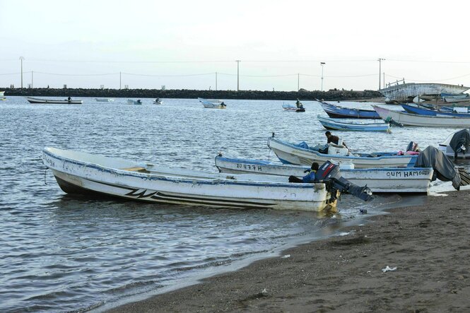 Einfache Boote mit Außenbordmotor liegen am Strand von Obock, in manchen Booten sitzt jeweils ein Mann, im Hintergrund ankern weitere Boote, die Hafenmauer begrenzt das Meer