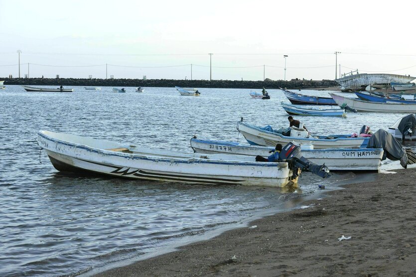 Einfache Boote mit Außenbordmotor liegen am Strand von Obock, in manchen Booten sitzt jeweils ein Mann, im Hintergrund ankern weitere Boote, die Hafenmauer begrenzt das Meer