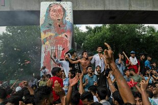 Protestierende feiern ausgelassen unter einem beschmierten Portrait der gerade gestürzten Premierministerin Sheikh Hasina