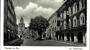 Straßenzug in Braunau 1935, das Haus auf der linken Seite, hat drei Stockwerke, Fensterbögen und Hakenkreuzfahnen hängen an der Fassade