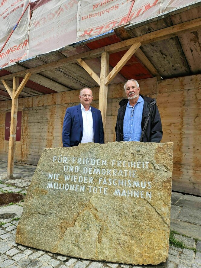 Zwei Männer mittleren Alters stehen vor der eingepackten Fassade von Hitlers Geburtsthaus, vor ihnen steht ein großer Gedenkstein mit der Inschrift: Für Frieden und Freiheit und Demokratie