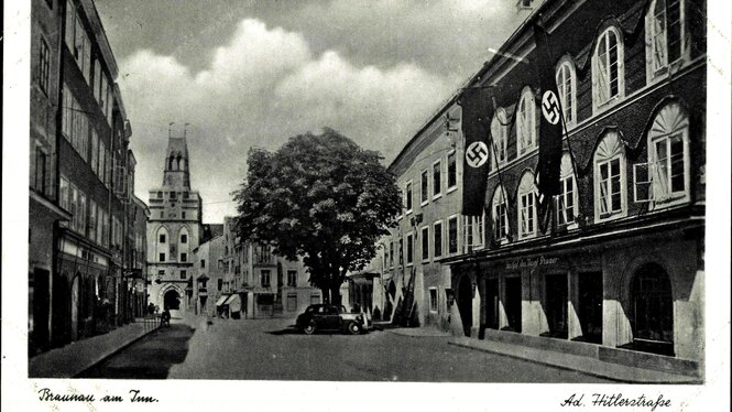 Straßenzug in Braunau 1935, das Haus auf der linken Seite, hat drei Stockwerke, Fensterbögen und Hakenkreuzfahnen hängen an der Fassade
