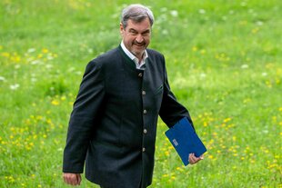 Markus Söder geht über eine grün-gelbe Wiese und hält einen blauen Ordner in der Hand