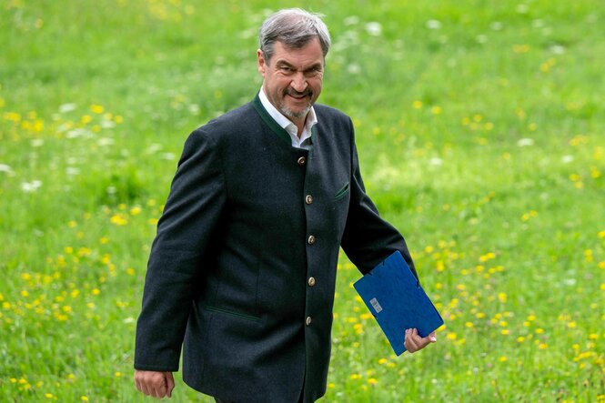 Markus Söder geht über eine grün-gelbe Wiese und hält einen blauen Ordner in der Hand