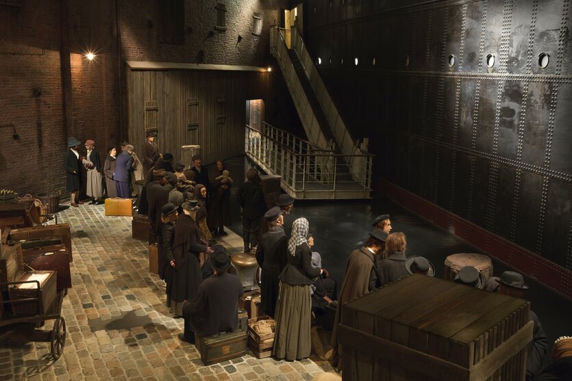 Menschen-Puppen in historischen Kleidern stehen in einer Schlange vor einem Schiff.