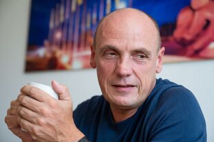Ein Porträtfoto des Politikers Daniel Born in Nahaufnahme. Er hält eine weiße Tasse in den Händen und blickt nachdenklich zur Seite, weg von der Kamera. Seine Lippen sind leicht geöffnet, was ihm einen konzentrierten und aufmerksamen Ausdruck verleiht, als würde er einem Gespräch folgen.