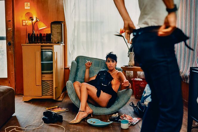 Eine leicht bekleidete Frau sitzt in einem Sessel und blickt auf eine Person, die sich gerade die Hose hochzieht.