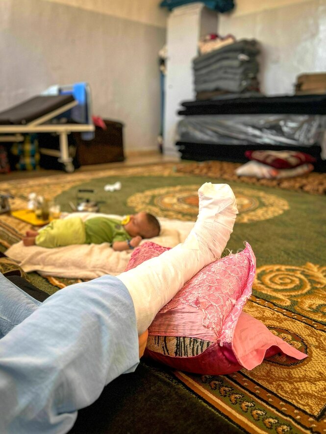 Ein Wohnzimmer, auf dem grün-gelben Teppich schläft ein Baby, im Vordergrund das eingewickelte Bein einer Person