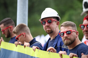 Männer in Arbeitskleidung halten ein Transparent, alle Männer trägen eine rote Brille auf der mit weißer Schrift steht: GAS KW JETZT!