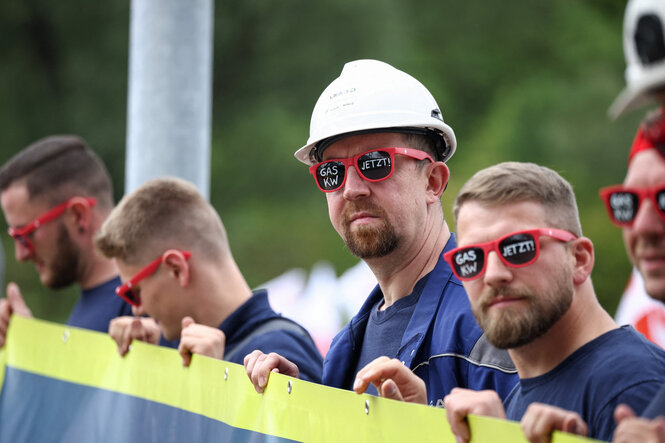 Männer in Arbeitskleidung halten ein Transparent, alle Männer trägen eine rote Brille auf der mit weißer Schrift steht: GAS KW JETZT!