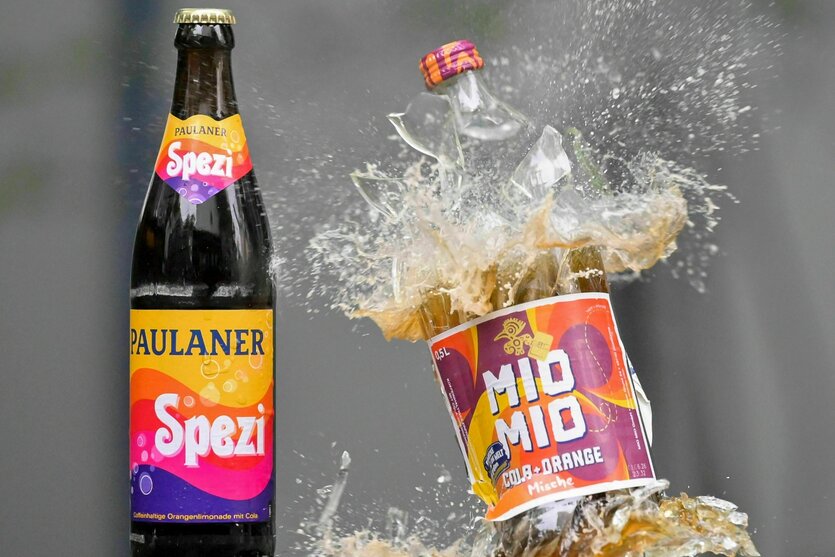 Eine Spezi-Flasche des Herstellers Paulaner (l) steht neben einer zerberstenden Flasche Mio-Mio-Cola-Orange-Mische des Herstellers Berentzen (r)